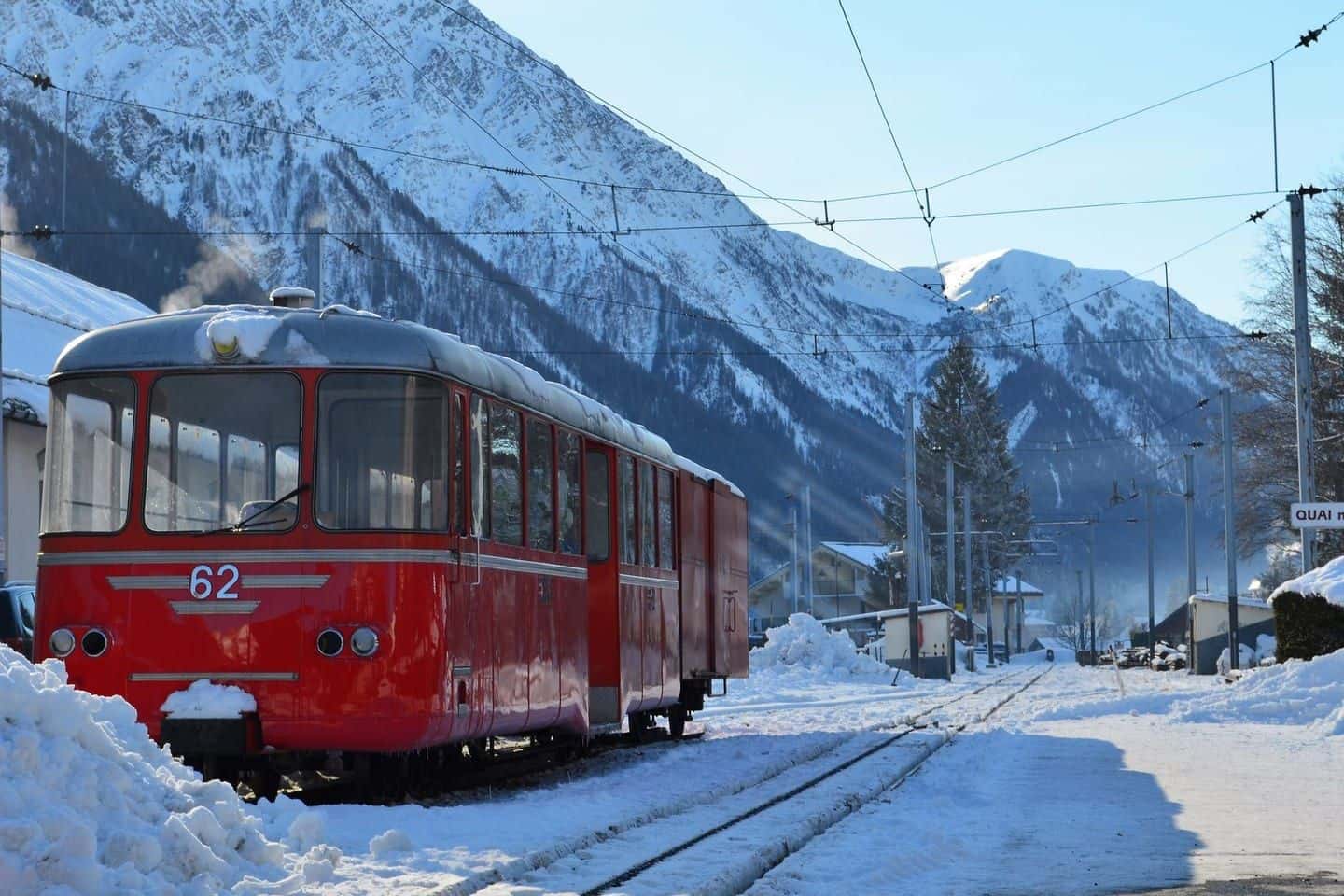 A red narrow-gauge mountain train waiting at a snowy station in the Chamonix valley, peaks behind