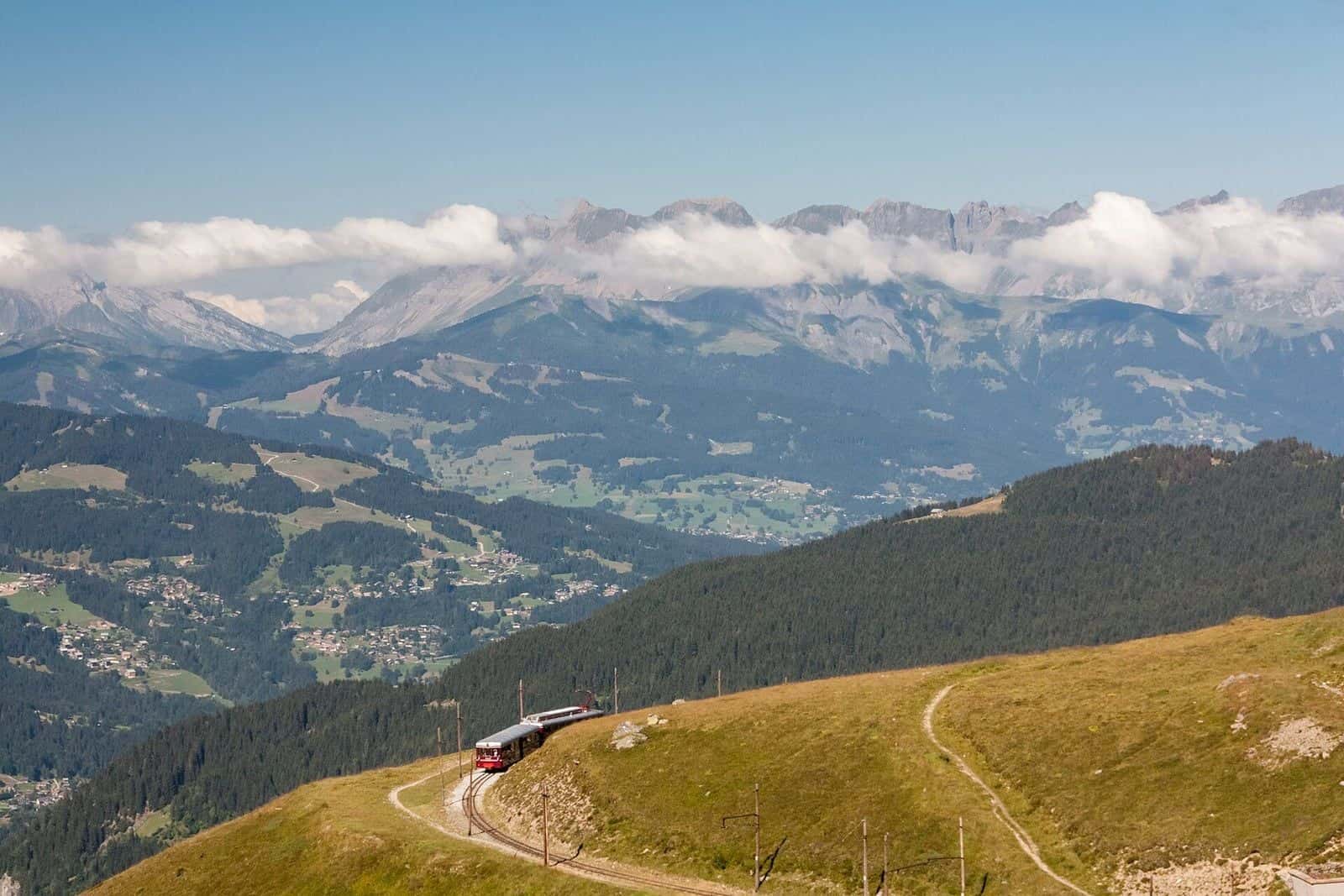 Rame du tramway du Mont-Blanc qui monte à travers les alpages au-dessus de Saint-Gervais, sommets enneigés et mer de nuages au loin